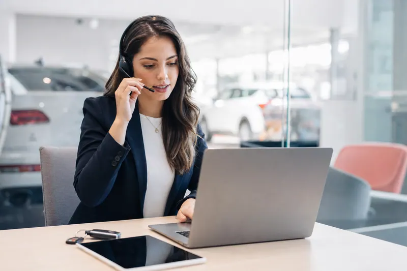 A dealership BDC rep on a headset call at a glass-walled desk